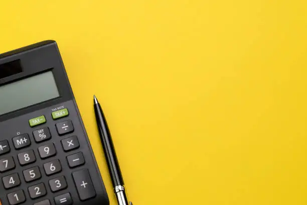 Photo of Flat lay or top view of black pen with calculator on vivid yellow background table with blank copy space, math, cost, tax or investment calculation