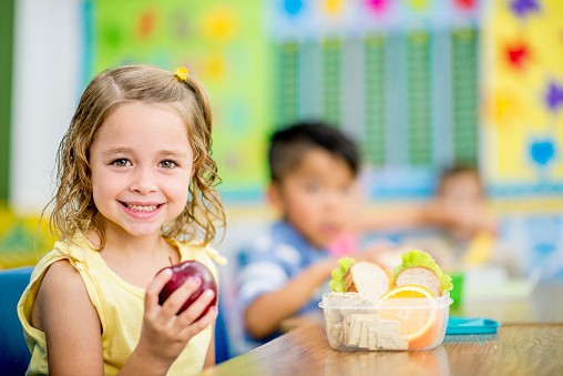 A young girl is sitting indoors in her elementary school classroom. She is smiling at the camera while holding up an apple.