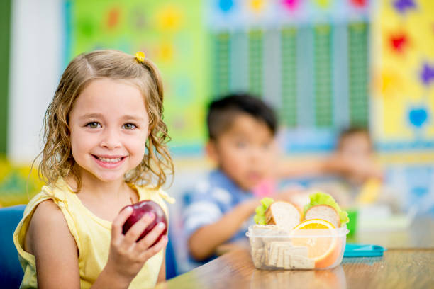 Eating An Apple A young girl is sitting indoors in her elementary school classroom. She is smiling at the camera while holding up an apple. school lunch child food lunch stock pictures, royalty-free photos & images