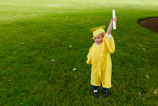Young, Caucasian boy is dressed in a yellow graduation gown, holding up his diploma with a green grass background