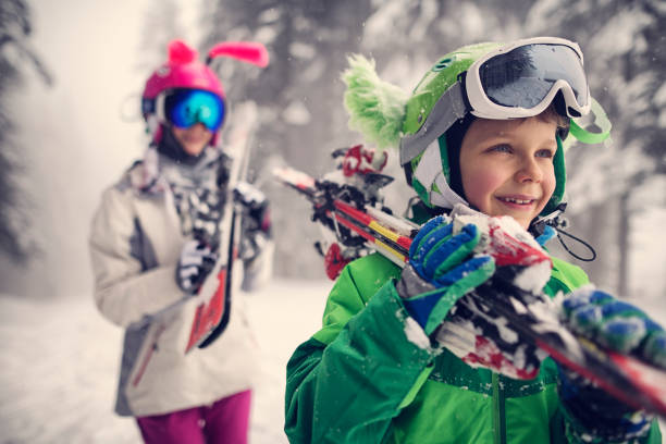 Little boy and his siter are carrying skis. Kids are wearing funny ski helmets. Beautiful winter day.\n