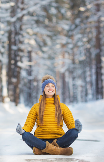 Young smiling woman with closed eyes in a bright clothes sits on a road (yoga) in a winter forest
