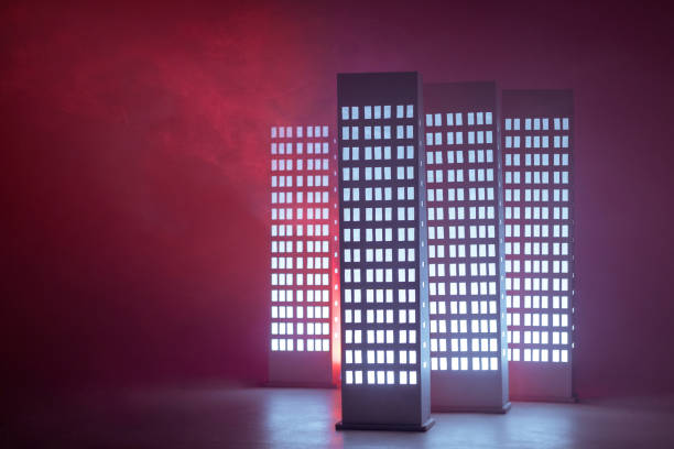A few cardboard models of high rise buildings with the rows of illuminating windows. The buildings maquettes (floor lamps) stands in different positions relative to each other. Studio shot on red background