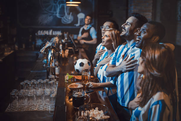 Happy soccer fans singing the anthem before the game in a bar. Group of happy sports fans singing the national anthem while watching the match on TV in a bar. Focus is on man with beard. national anthem stock pictures, royalty-free photos & images