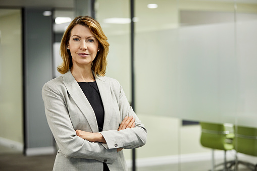 Portrait of confident businesswoman standing against board room. Blond entrepreneur is with arms crossed in lobby at office. She is wearing businesswear.