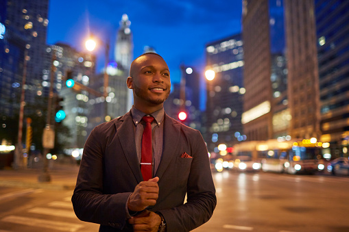 Confident businessman buttoning cuff in illuminated city. Mid adult professional is smiling while looking away. He is wearing suit on street.