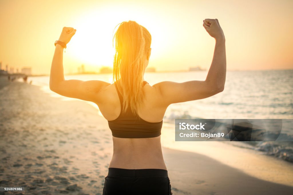 Vue arrière du forte fille sportive montrant les muscles à la plage pendant le coucher du soleil. - Photo de Plage libre de droits