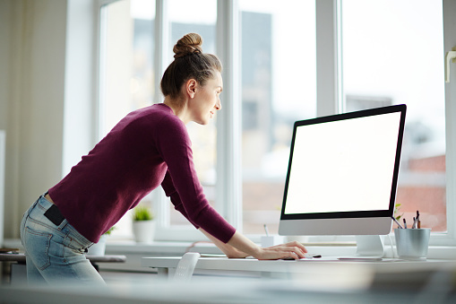 Young programmer or web-designer in purple pullover and blue jeans leaning over desk while reading online information on monitor display