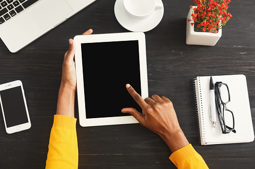 Black female hands on digital tablet with blank screen for advertisement. Top view of african-american hands, laptop keyboard, coffee, smartphone on a wooden office table background.