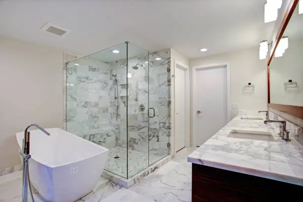 Sleek bathroom features freestanding bathtub paired with floor-mounted faucet atop marble floor placed in front of glass shower accented with rain shower head and gray marble surround.