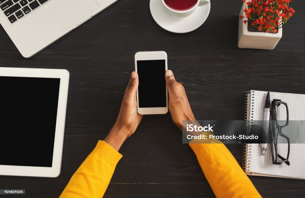 Black female hands holding smartphone, top view Black female hands holding smartphone with blank screen for advertisement. Top view of african-american hands, laptop keyboard, coffee, mobile on dark wooden table background, copy space Dark Stock Photo