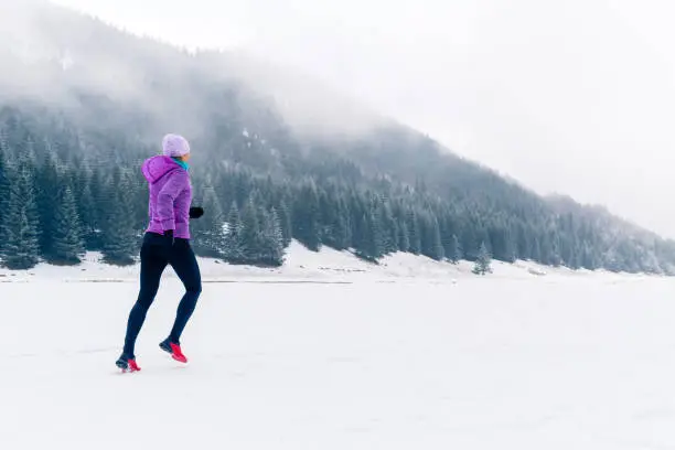 Woman trail running in mountains on snow footpath, winter day. Sport, fitness inspiration and motivation. Girl cross country runner training in cold conditions.