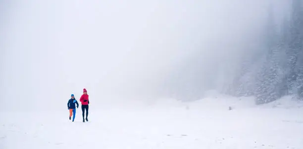 Girls running together on snow in winter mountains. Sport, fitness inspiration and motivation. Two women partners trail running in mountains, winter day. Female trail runner jogging outdoors.