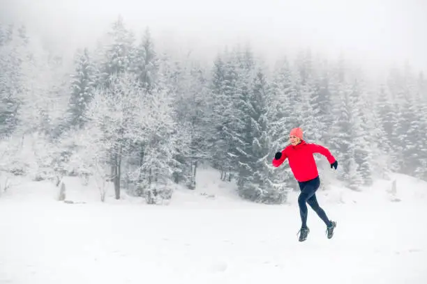 Woman trail running on snow in winter mountains and forest. Sport, fitness inspiration and motivation. Young happy girl athlete working out on snow, winter day. Female runner jogging outdoors.