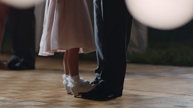 Young girl dances on her father's feet under twinkling lights at wedding reception.