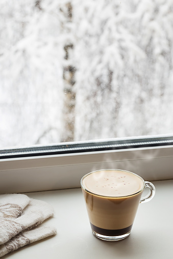 Cozy winter still life: cup of hot coffee and warm mittens on windowsill against the background of snow-covered trees outside the window