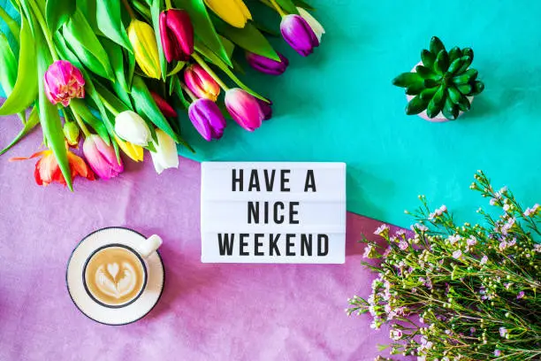 Shot from above as flatlay with Have a nice weekend message written on light box with coffee and colorful tulips and flowers