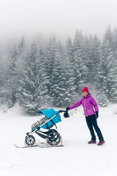 Mother with baby stroller enjoying motherhood in winter forest, mountains landscape. Jogging or power walking woman with sledge pram in woods. Beautiful winter snowy inspirational mountains.