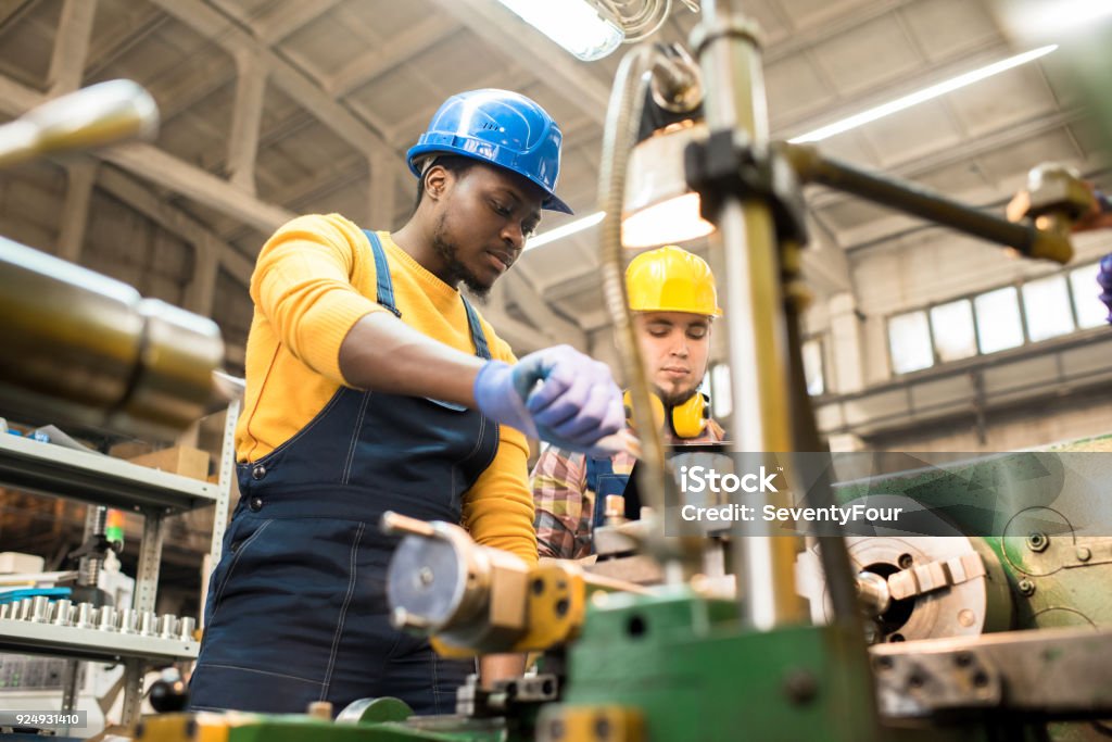 Lathe Operators Concentrated on Work Multi-ethnic team of workers wearing overalls and protective helmets using lathe in order to machine workpiece, interior of spacious production department on background Manufacturing Stock Photo