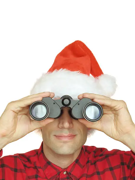 This color photo shows a smiling man with a fuzzy red Santa hat looking through black binoculars directly at the viewer. He is visible from the shoulders up, wearing a black and red flannel shirt, and holding binoculars with both hands. The image is isolated on a white background in portrait orientation.