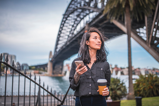 Mid adult Asian woman is using phone on footpath in Sydney, Australia. Bridge in the back.