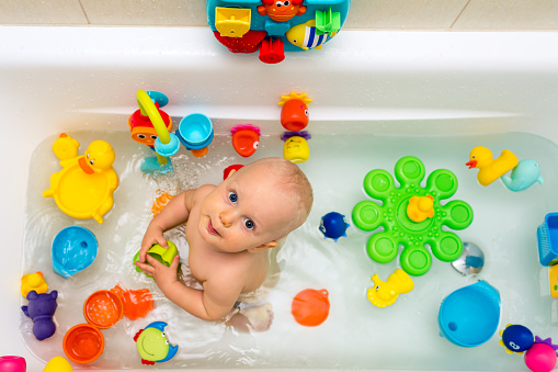 Baby boy taking a bath, playing in a bathtub with colorful toys. Smiling little kid in bathroom looking at camera. Infant playing, have fun and washing. Healthy hygiene and care for children.