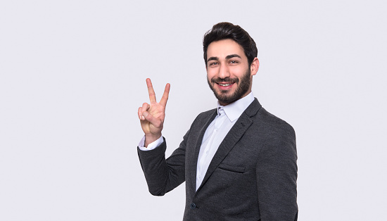 Portrait of smiling young man gesturing peace sign against gray background. Horizontal composition. Image taken with Nikon D800 and developed from Raw format. Studio shot.