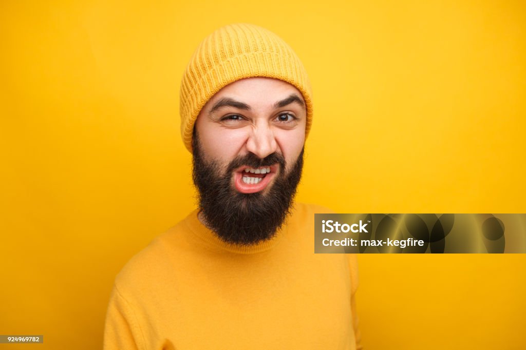 Grimacing man in yellow looking at camera Bearded hipster man in yellow hat and sweater grimacing at camera on yellow backdrop. Adult Stock Photo