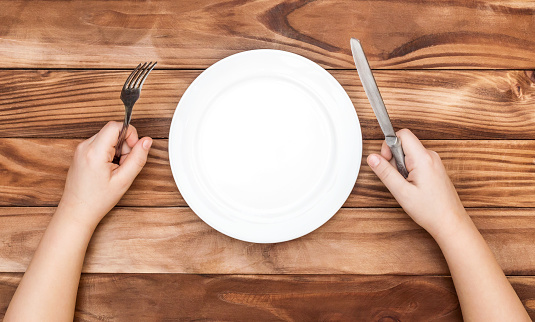 Hungry child waiting for meal. Child's hands holding fork and knife over table with empty dish. Top view.