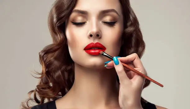 Graceful hands of professional make-up master, with the blue manicure on the nails,  is painting in the red color lips of splendid young woman. Creation of perfect make up in progress. Makeup,manicure and cosmetic.