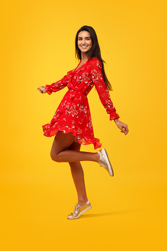 Bright charming female in red flying dress dancing on orange background smiling at camera.