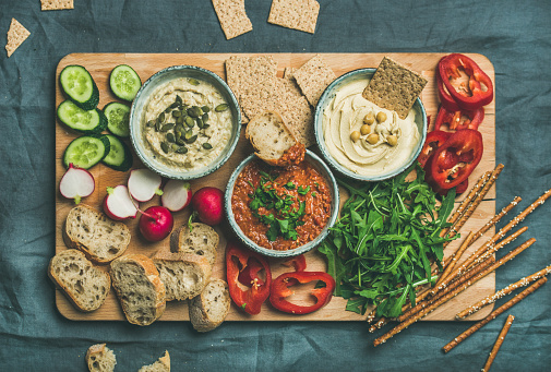 Vegan snack board. Flat-lay of Various Vegetarian dips hummus, babaganush and muhammara with crackers, bread, fresh vegetables on wooden board over grey background. Clean eating, dieting food concept