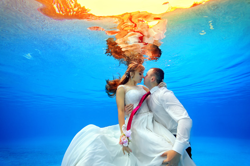 Couple in wedding dresses hugging and kissing underwater in the pool at sunset. Horizontal view. Shooting from under the water
