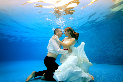 Happy girl and man in wedding dresses underwater hugging the bottom of the pool and looking at each other. Portrait. Landscape orientation. Shooting under water