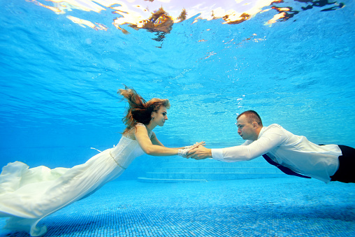 The bride and groom in wedding dresses swim underwater in the pool to meet each other. Portrait. Landscape orientation. Shooting under water