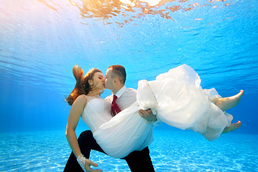 The happy groom holds the bride in his arms underwater in the pool and kisses her on the background of sunlight. Portrait. Shooting under water. Landscape orientation