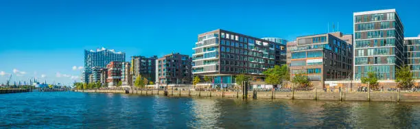 Panoramic view across the blue waters of the Norderelbe to the crowded promenade and modern apartment buildings of the redeveloped HafenCity quarter of Hamburg, Germany.