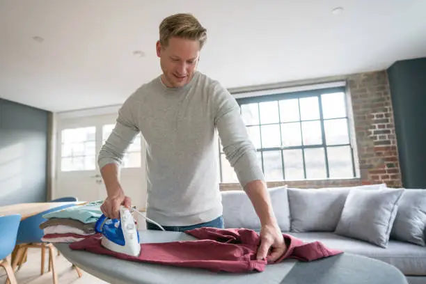Handsome single guy at home ironing his clothes looking happy and smiling