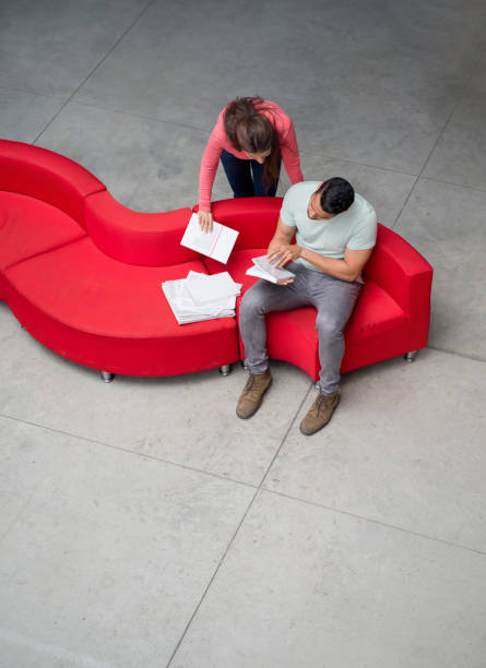 istock Young man showing her colleague a book he is reading while sitting on a red couch 925033012