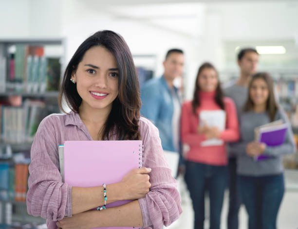 istock Latin american female student at the library holding a notebook lookign at camera smiling 925033046