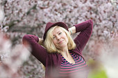 Caucasian woman with long blond hair and purple fedora hat near blossoming tree, arms behind her head with the elbows pointing out