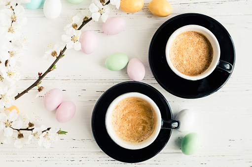 Two cups of coffee espresso, colorful chocolate eggs and cherry blossom on white shabby chic background. Easter treats concept. Top view.