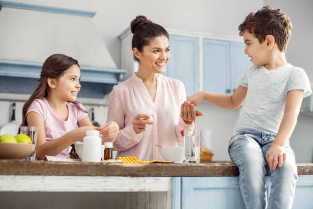 Delighted mother having breakfast with her children Happy mother. Attractive alert dark-haired young mother smiling and giving pills to her son sitting on the table and her daughter having breakfast vitamin stock pictures, royalty-free photos & images