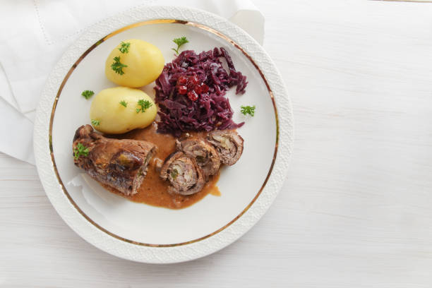 istock beef roll with red cabbage and potatoes, german meat roulade stuffed with cucumbers, bacon and onions on an elegant gold rim plate on a white wooden table, high angle view from above, copy space 925072616