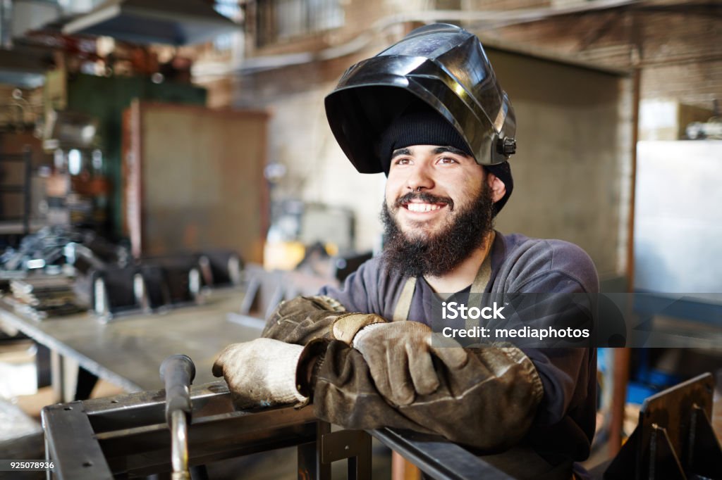 Happy excited welder enjoying manual work in factory shop Cheerful dreamy bearded young man with dirty sleeves wearing welding mask and looking with burning eyes while leaning on metal part in factory shop Welder Stock Photo