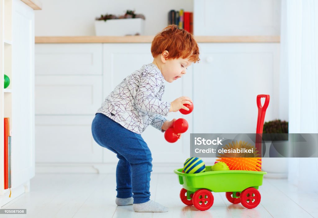 cute redhead toddler baby collecting different balls into toy pushcart Child Stock Photo