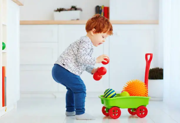 Photo of cute redhead toddler baby collecting different balls into toy pushcart