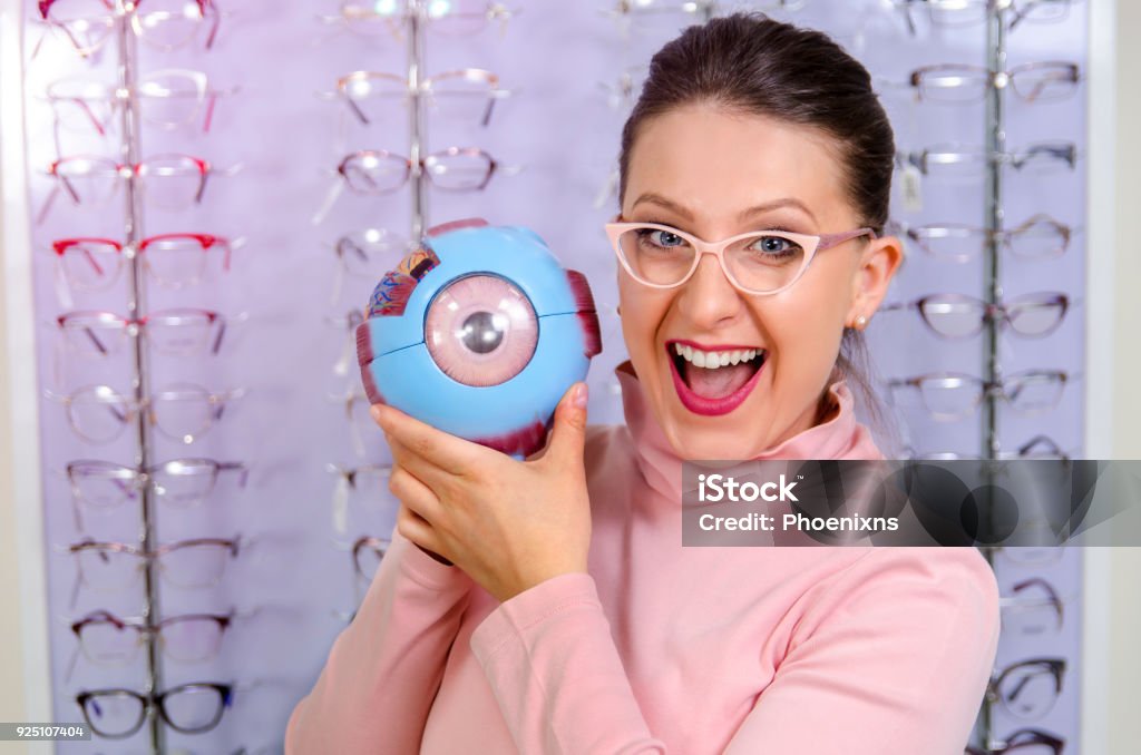 Young woman in optics store with plastic eye model Cute young woman in optics store holding plastic eye model and smiling , optometry concept Adult Stock Photo