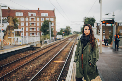 Vintage toned portrait of a young beautiful brunette woman, standing on the train station, waiting for her public transportation. She is wearing a casual outfit, an olive green parka jacket.