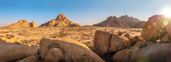 Wide angle view of the dusty, reddish brown rocky, rocky landscape and the ancient geological formations of Namibia's Spitzkoppe. Sun rising with lens flare over a large granite boulder, a dirt road visible. Blue sky in the background.
